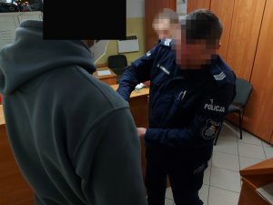 Na zdjęciu widać umundurowanego policjanta, który zakłada kajdanki na ręce mężczyzny. Zdjęcie jest wykonane w pomieszczeniu służbowym.