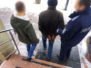 Na zdjęciu widać dwóch policjantów, którzy pomiędzy sobą prowadzą mężczyznę. Mężczyzna ma założone kajdanki na ręce trzymane z tyłu. Zdjęcie jest wykonane na schodach przed drzwiami wejściowymi do budynku komendy. Wszystkie osoby są odwrócone tyłem do zdjęcia.