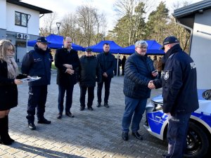 Na zdjęciu wdać przedstawicieli z Gminy Wiązowna, którzy w asyście Komendanta Powiatowego Policji w Otwocku przekazują policjantowi stojącemu przy oznakowanym radiowozie kluczyki.