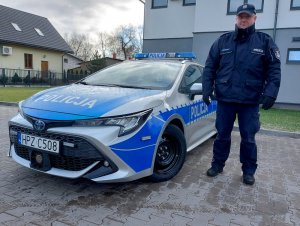 Na zdjęciu widać oznakowany radiowóz, obok którego stoi umundurowany policjant.
