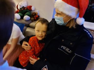 Na zdjęciu widać umundurowaną policjantkę z czapką Mikołaja, która przytula chłopca. Zdjęcie jest wykonane w pomieszczeniu Domu dla Dzieci.