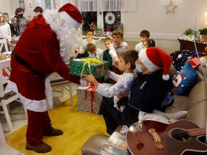 Na zdjęciu widać Mikołaja, który wręcza dzieciom prezenty. Zdjęcie jest wykonane w pomieszczeniu w Domu dla Dzieci.