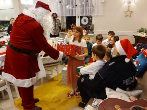 Na zdjęciu widać Mikołaja, który wręcza dzieciom prezenty. Zdjęcie jest wykonane w pomieszczeniu w Domu dla Dzieci.