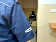 Na zdjęciu widać bark umundurowanego policjanta, w tle widać siedzącego na łóżku w policyjnej celi mężczyznę.