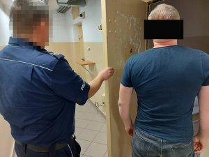 Na zdjęciu widać umundurowanego policjanta, który trzyma otwarte drzwi do policyjnej celi. Obok niego stoi mężczyzna, odwrócony tyłem do zdjęcia.