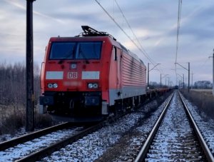 Na zdjęciu widać stojący na torach pociąg koloru czerwonego. Zdjęcie jest wykonane z lewej strony lokomotywy.