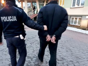Na zdjęciu widać umundurowanego policjanta z drogówki, który prowadzi mężczyznę z założonymi kajdankami na ręce trzymane z tyłu w kierunku komendy.
