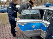 Na zdjęciu widać umundurowanego policjanta, który trzyma otwarte drzwi oznakowanego radiowozu. Przed nim mężczyzna siada na tylną kanapę pojazdu.