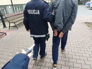 Na zdjęciu widać umundurowanego policjanta, który prowadzi mężczyznę z założonymi kajdankami na ręce trzymane  z tyłu. Obaj są odwróceni tyłem do zdjęcia. Zdjęcie jest wykonane przed budynkiem komendy.