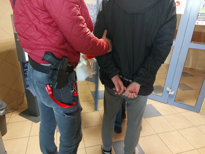 Na zdjęciu widać nieumundurowanego policjanta, który trzyma mężczyznę z założonymi kajdankami na ręce trzymane z tyłu. Zdjęcie jest wykonane na korytarzu budynku komendy.