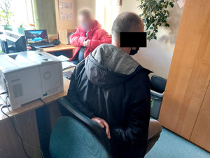 Na zdjęciu widać siedzącego na krześle mężczyznę z założonymi kajdankami na ręce trzymane  z tyłu. Po drugiej stronie biurka siedzi nieumundurowany policjant. Na blacie znajduje się komputer i dokumenty. Zdjęcie jest wykonane w pomieszczeniu służbowym.