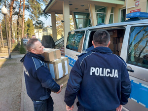 Na zdjęciu widać umundurowanych policjantów, którzy pakują kartony do oznakowanego radiowozu typu bus.