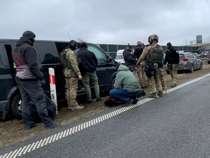 Na zdjęciu widać dziewięciu funkcjonariuszy pionu kryminalnego i jednostek specjalnych, w tle nieoznakowane radiowozy. Jeden z nieumundurowanych policjantów trzyma leżącego na ziemi mężczyznę. Zdjęcie jest wykonane przy autostradzie.