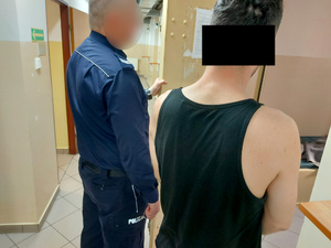 Na zdjęciu widać umundurowanego policjanta, obok którego stoi mężczyzna. Obaj są odwróceni tyłem do zdjęcia. Zdjęcie jest wykonane przed drzwiami do wejściowymi do policyjnej celi.