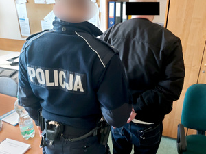 Na zdjęciu widać umundurowanego policjanta, który trzyma mężczyznę z założonymi kajdankami na ręce trzymane z tyłu. Obaj są odwróceni tyłem do zdjęcia. Zdjęcie jest wykonane w pomieszczeniu służbowym.