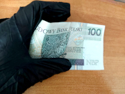 Na zdjęciu jest dłoń ubrana w czarną rękawicę, która trzyma banknot o nominale 100 złotych.