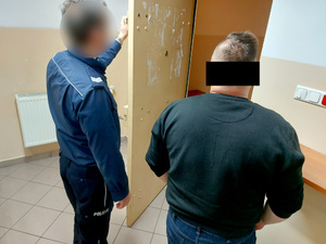 Na zdjęciu widać umundurowanego policjanta, który trzyma otwarte drzwi do policyjnej celi. Obok niego stoi mężczyzna. Obaj są wróceni tyłem do zdjęcia.