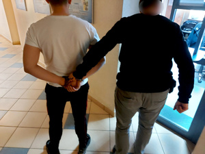 Na zdjęciu widać nieumundurowanego policjanta, który trzyma mężczyznę z założonymi kajdankami na ręce trzymane z tyłu. Obaj są odwróceni tyłem do zdjęcia. Zdjęcie jest wykonane na korytarzu budynku jednostki.