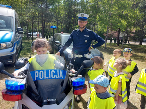 Dziewczynka siedząca na policyjnym motocyklu. Obok motocykla stoją dzieci i umundurowany policjant.