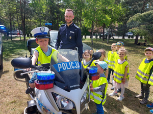 Chłopiec siedzący na policyjnym motocyklu. Obok motocykla stoją dzieci i umundurowany policjant.