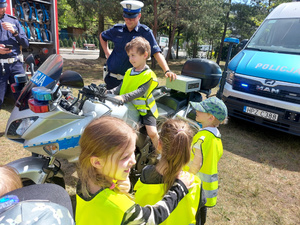 Chłopiec siedzący na policyjnym motocyklu. Z prawej strony motocykla stoi umundurowany policjant, z lewej dzieci.
