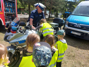 Na zdjęciu widać umundurowanego policjant, który pomaga wsiąść chłopcu na policyjny motocykl. Obok motocykla stoją dzieci.