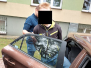 Na zdjęciu widać nieumundurowany policjanta, który trzyma otwarte, tylne drzwi nieoznakowanego radiowozu. Przed nim jest mężczyzna, który wsiada na tylną kanapę pojazdu. Obaj są odwróceni przodem do zdjęcia.