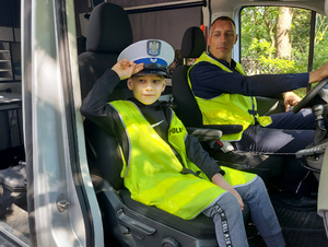 Chłopiec w policyjnej czapce i odblaskowej kamizelce siedzący na przednim siedzeniu pasażera w oznakowanym radiowozie typu bus. Za kierownicą pojazdu siedzi umundurowany policjant.