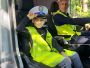 Chłopiec w policyjnej czapce i odblaskowej kamizelce siedzący na przednim siedzeniu pasażera w oznakowanym radiowozie typu bus. Za kierownicą pojazdu siedzi umundurowany policjant.