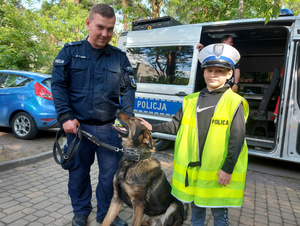 Przewodnik z psem służbowym, obok którego stoi chłopiec ubrany w policyjną, białą czapkę i odblaskową kamizelkę.