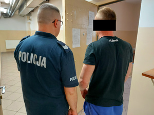 Na zdjęciu widać umundurowanego policjanta, który trzyma otwarte drzwi do policyjnej celi. Obok niego stoi mężczyzna. Obaj są odwróceni tyłem do zdjęcia.