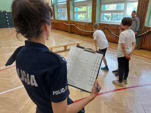 Zdjęcie stojącej tyłem umundurowanej policjantki, która trzyma w ręku tabelę punktową. W tle widać uczestników konkursu w trakcie pokonywania toru przeszkód.