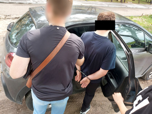 Na zdjęciu widać nieumundurowanego policjanta, który trzyma mężczyznę z założonymi kajdankami na ręce trzymane z tyłu. Obaj stoją przed otwartymi, tylnymi drzwiami nieoznakowanego radiowozu.