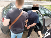 Na zdjęciu widać nieumundurowanego policjanta, który trzyma mężczyznę z założonymi kajdankami na ręce trzymane z tyłu. Obaj stoją przed otwartymi, tylnymi drzwiami nieoznakowanego radiowozu.