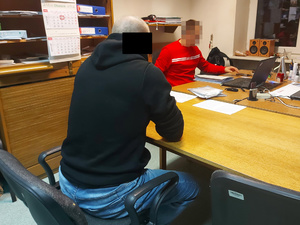 Na zdjęciu widać mężczyznę, który siedzi przy biurku. po drugiej stronie siedzi nieumundurowany policjant. Na blacie jest komputer i dokumenty.