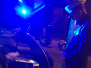 Na zdjęciu widać umundurowanego policjanta z drogówki, który stoi przy otwartym bagażniku radiowozu, gdzie znajduje się urządzenie do badania trzeźwości. Zdjęcie jest wykonane porą nocną.