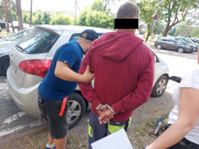 Na zdjęciu widać dwóch policjantów, którzy trzymają pomiędzy sobą mężczyznę. Mężczyzna ten ma założone kajdanki na ręce trzymane z tyłu. Zdjęcie jest zrobione tuż przez nieoznakowanym radiowozem.