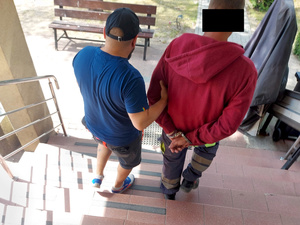 Na zdjęciu widać policjanta, który trzyma mężczyznę. Mężczyzna ma założone kajdanki na ręce trzymane z tyłu. obaj są odwróceni tyłem do zdjęcia. Zdjęcie jest wykonane na schodach przed drzwiami wejściowymi do budynku komendy.